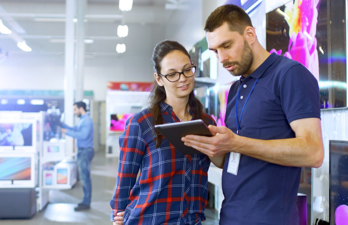 In The Electronics Store Professional Consultant Provides Expert Advice On Tablet Computer Specifications For Beautiful Young Woman. Store Is Bright, Modern And Has All The Latest Devices.